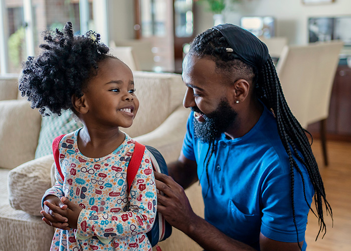 Young girl smiles at her father as he helps her put on a backpack.