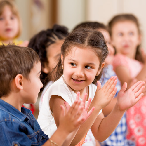 A young child smiles at another preschooler as they and their classmates clap hands, links to information on making friends in early childhood.