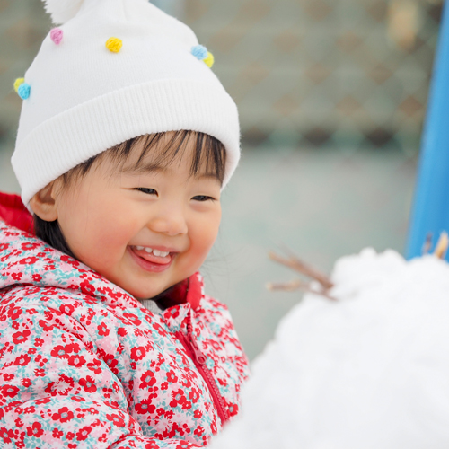 A toddler in winter hat and coat smiles at a snowman, links to information on play in all types of weather.